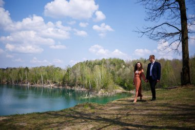 Mutlu ve genç evliler el ele tutuşup gülüyorlar. Gölün arka planında ve yeşil bir çayırda. Damat ve kıvırcık saçlı güzel gelin çayırda yürüyorlar. Neşeli bir yaz günü