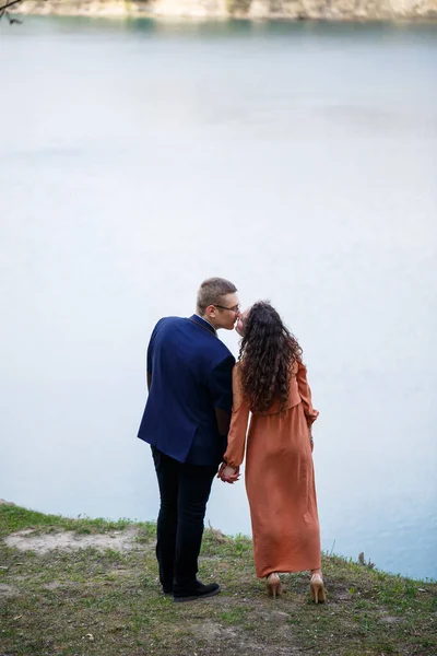 青い湖を背景に手をつないでいる若いカップル 長いドレスの女の子と自然の上にスーツの男 ロマンスと愛幸せなカップル — ストック写真