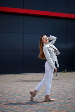 Avrupa görünümlü uzun sarı saçlı, yüzünde bir gülümsemeyle şık bir genç kadın. Beyaz ceketli ve beyaz kotlu bir kız. Gri bir binanın arka planında güneşli sıcak bir yaz günü.