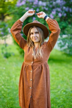 Sarı saçlı güzel bir kız elinde bir şapka tutuyor ve gözleri leylak çalılarının yanında dikiliyor. Çiçek açan ağaçları olan bir bahçede genç bir kadın