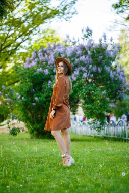 Kahverengi şapkalı şık bir kız ve eflatun renkli çalıların üzerinde açık renk bir elbise. Güneşli bir yaz gününde yüzünde bir gülümsemeyle parkta yürüyen genç bir kadın.