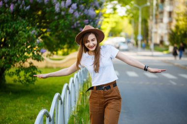 Kahverengi şapkalı ve beyaz tişörtlü güzel bir kız sıcak ve güneşli bir günde yolda yürüyor. Yüzünde bir gülümseme olan şapkalı güzel bir genç kadın.