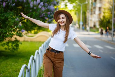 Kahverengi şapkalı ve beyaz tişörtlü güzel bir kız sıcak ve güneşli bir günde yolda yürüyor. Yüzünde bir gülümseme olan şapkalı güzel bir genç kadın.