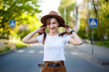 Kahverengi şapkalı ve beyaz tişörtlü güzel bir kız sıcak ve güneşli bir günde yolda yürüyor. Yüzünde bir gülümseme olan şapkalı güzel bir genç kadın.