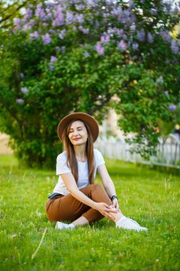 Parktaki yeşil çimlerde oturan şapkalı mutlu genç bir kadın. Güneşli bir yaz gününde yüzünde gülümseme olan Avrupalı bir kız.