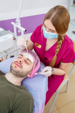Kozmetik uzmanı, güzellik kliniğinde fırça kullanarak adamın yüzüne beyaz nemlendirici maske takıyor. Güzellik endüstrisi konsepti. Adam kanepede yatıyor. Güzellik uzmanı kadın spa prosedürü yapıyor..