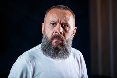 Beyaz tişörtlü, sakallı, yetişkin, gri saçlı bir adamın duygusal portresi.