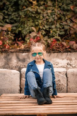 Küçük tatlı bir kız güneş gözlükleriyle taş bir çitte oturuyor. Mavi ceketli şık bir çocuk. Sıradan tarz, çocuklar için moda, şık takım elbise, mutlu çocukluk.