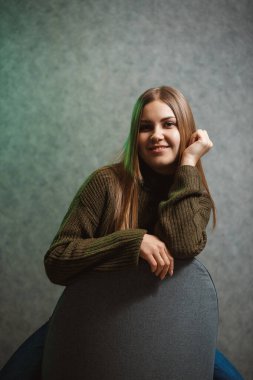 Yeşil kazaklı, kot pantolonlu, gri sandalyeli ve gülümseyen bir kız.