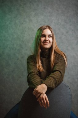 Yeşil kazaklı, kot pantolonlu, gri sandalyeli ve gülümseyen bir kız.