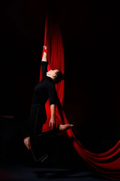 A sports girl performs gymnastic and circus exercises on red silk. Studio shooting on a dark background. Aerial gymnastics on canvas