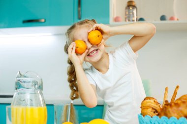 7-8 yaşlarında bir kız mutfakta oturur ve mandalinayla göz yapar..