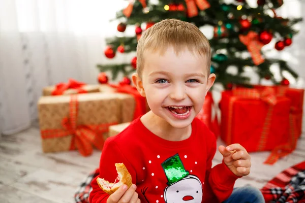 Le garçon assis près du sapin de Noël pour la nouvelle année. Décor de Noël avec des cadeaux, un enfant près du sapin de Noël mange des croissants et sourit — Photo