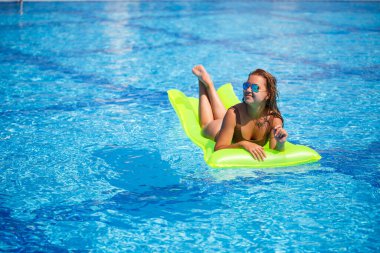 Havuzda şişme bir şiltenin üzerinde mayo giymiş genç seksi bir kadın yüzüyor. Tatilde mavi havuzda güneş gözlüklü bir kız.