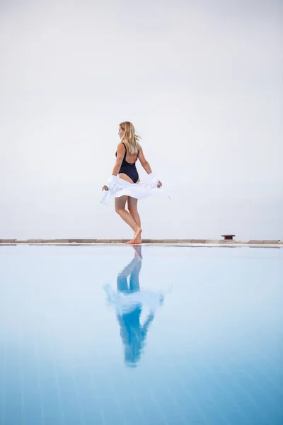 Een aantrekkelijke jonge vrouw in een zwart badpak en wit shirt staat bij het zwembad met een panoramisch uitzicht. — Stockfoto