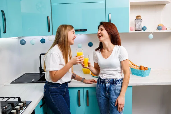 Two women friends standing in the kitchen and drinking orange juice. Girlfriends chat and share secrets in the kitchen, breakfast