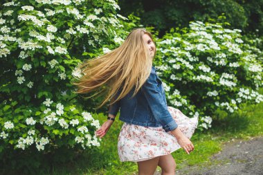 Avrupa görünümlü, uzun sarı saçlı, kısa bir elbise giymiş genç bir kadın, beyaz çiçekli çalıların arka planında duruyor. Güneşli bir bahar günü. Doğal kadın güzelliği