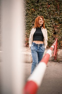 Güzel kızıl saçlı genç bir kadın sokakta yürüyor, kot pantolon ve bej bir gömlek giyiyor. Yüzünde bir gülümsemeyle sıradan giyinmiş güzel bir kız.