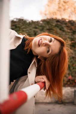 Güzel kızıl saçlı genç bir kadın sokakta yürüyor, kot pantolon ve bej bir gömlek giyiyor. Yüzünde bir gülümsemeyle sıradan giyinmiş güzel bir kız.