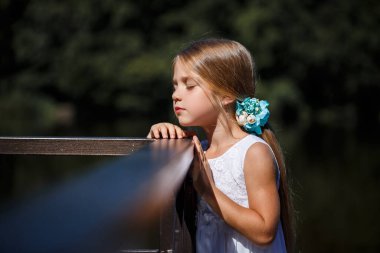 6-7 yaşlarında, uzun sarı saçlı, bale elbisesi giymiş, nehrin arka planında duran tatlı bir kız çocuğu.