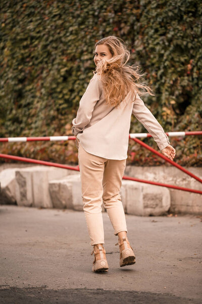 A pretty blonde young woman is walking down the street, she is wearing jeans and a beige shirt. Beautiful girl dressed in casual style with a smile on her face for a walk