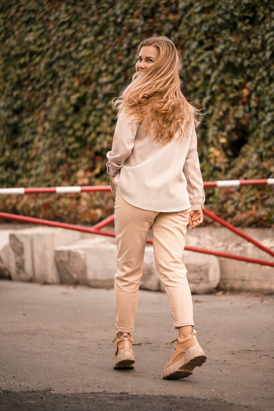 A pretty blonde young woman is walking down the street, she is wearing jeans and a beige shirt. Beautiful girl dressed in casual style with a smile on her face for a walk