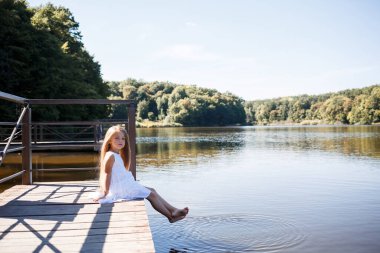 Göl kenarındaki uzun sarı saçlı 7 yaşında bir kız suda bacaklarıyla debriyajda oturuyor. Ayaklarını göle atıyor. Uzun saçlı, beyaz elbiseli çıplak ayaklı kız..