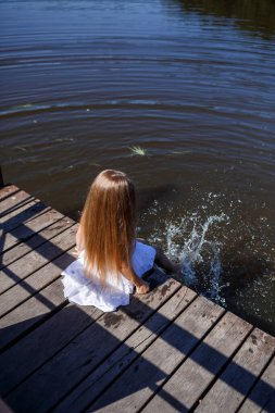 Göl kenarındaki uzun sarı saçlı 7 yaşında bir kız suda bacaklarıyla debriyajda oturuyor. Ayaklarını göle atıyor. Uzun saçlı, beyaz elbiseli çıplak ayaklı kız..