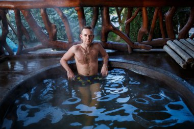 Terasta sıcak suyla havuzda genç bir adam, spa vücut bakımı. Açık hava jakuzisi. Bir adamla jakuzi. Açık havada rahatla. Kaplıca banyosu olan bir kulübe..