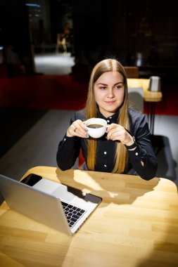 Elinde bir fincan kahve tutan ve dizüstü bilgisayar kullanan genç bir kadın. Evden çalışan bir iş kadını. Çevrimiçi alışveriş, e-ticaret, evden çalışma