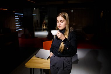 Modern kadın işadamının normal çalışma günü. Güzel genç bir kadın iş yerinde otururken elinde bir fincan kahve tutuyor.