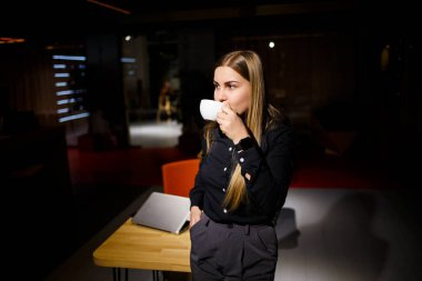 Modern kadın işadamının normal çalışma günü. Güzel genç bir kadın iş yerinde otururken elinde bir fincan kahve tutuyor.