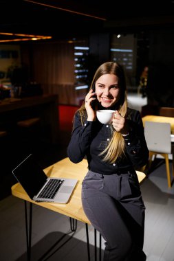 Elinde bir fincan kahve tutan ve dizüstü bilgisayar kullanan genç bir kadın telefonda konuşuyor. Evden çalışan bir iş kadını. Evden çalış.