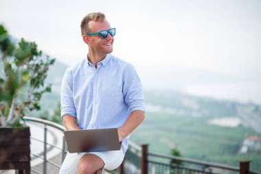 Genç başarılı erkek işadamı deniz kenarında dizüstü bilgisayarla çalışıyor. Güneş gözlüğü, gömlek ve beyaz şort giyiyor. Ofisin dışında çalış, serbest çalışan.