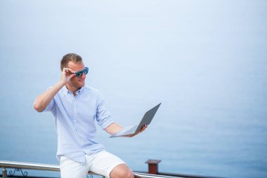 Genç başarılı erkek işadamı deniz kenarında dizüstü bilgisayarla çalışıyor. Güneş gözlüğü, gömlek ve beyaz şort giyiyor. Ofisin dışında çalış, serbest çalışan.