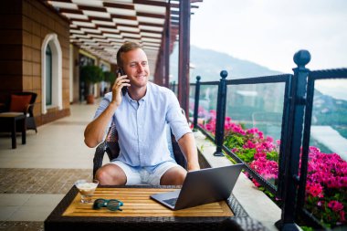 Beyaz gömlekli ve şortlu genç yakışıklı bir işadamı masada dizüstü bilgisayarla oturuyor ve telefonla konuşuyor. Dinlenirken çalış