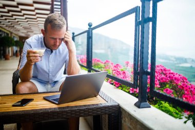 Serbest yazar konsepti. Başarılı genç adam, iş adamı dizüstü bilgisayarda çalışıyor, terasta oturuyor kahve içiyor ve telefonda konuşuyor..