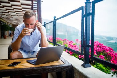 Serbest yazar konsepti. Başarılı genç adam, iş adamı dizüstü bilgisayarda çalışıyor, terasta oturuyor kahve içiyor ve telefonda konuşuyor..