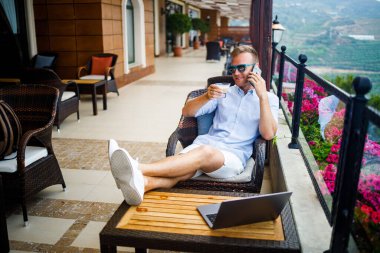 Serbest yazar konsepti. Başarılı genç adam, iş adamı dizüstü bilgisayarda çalışıyor, terasta oturuyor kahve içiyor ve telefonda konuşuyor..