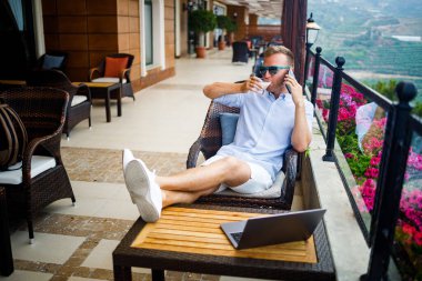 Serbest yazar konsepti. Başarılı genç adam, iş adamı dizüstü bilgisayarda çalışıyor, terasta oturuyor kahve içiyor ve telefonda konuşuyor..