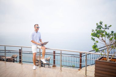 Dizüstü bilgisayarla çalışan yakışıklı ve başarılı genç iş adamı Akdeniz 'e bakıyor. O bir gömlek ve beyaz şort giyiyor. Tatilde uzaktan çalışma. Tatil kavramı