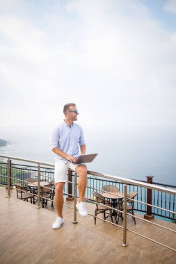 Dizüstü bilgisayarla çalışan yakışıklı ve başarılı genç iş adamı Akdeniz 'e bakıyor. O bir gömlek ve beyaz şort giyiyor. Tatilde uzaktan çalışma. Tatil kavramı