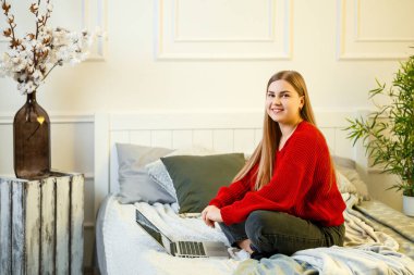 Genç bir kadın bilgisayarda çalışıyor, yatakta oturuyor, uzaktan çalışıyor. Kırmızı kazaklı ve kot pantolonlu uzun saçlı bir kız evde çalışıyor..