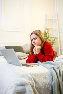 Genç bir kadın bilgisayarda çalışıyor, yatakta oturuyor, uzaktan çalışıyor. Kırmızı kazaklı ve kot pantolonlu uzun saçlı bir kız evde çalışıyor..