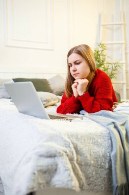 Kırmızı kazaklı genç bir kadın parlak bir odada dizüstü bilgisayarda oturuyor. Karantina sırasında ev atmosferinde uzaktan çalışma