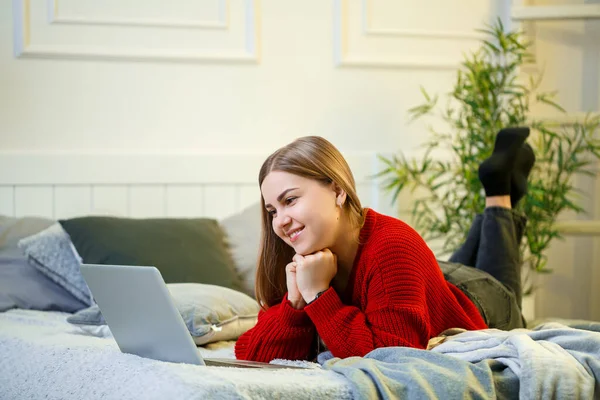 Young woman works at a computer, sitting on a bed, working from a distance. A girl with long hair in a red sweater and jeans works at home.