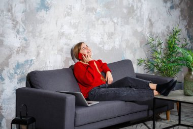 Güzel ve çekici genç bir kadın evinde kanepede oturuyor ve telefonda konuşuyor. Kırmızı bir kazak ve kot pantolon giyiyor, dizüstü bilgisayarın yanında.