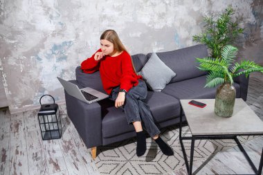 Kanepede oturan genç bir kadın dizüstü bilgisayarın ekranına bakıyor. İnternette bilgisayar üzerinde çalışan motive olmuş, ev tabanlı, serbest çalışan bir bayan öğrenci..