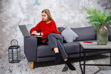 Kanepede oturan genç bir kadın dizüstü bilgisayarın ekranına bakıyor. İnternette bilgisayar üzerinde çalışan motive olmuş, ev tabanlı, serbest çalışan bir bayan öğrenci..