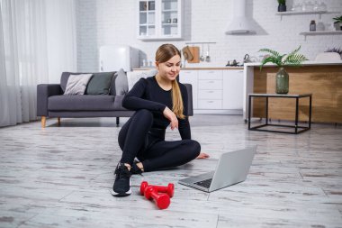 Siyah bluzlu ve taytlı genç bir bayan antrenör dizüstü bilgisayarla oturur ve karantina sırasında evde spor ve spor programı izler. Sağlıklı bir yaşam tarzı. Dambıllar yere düştü.